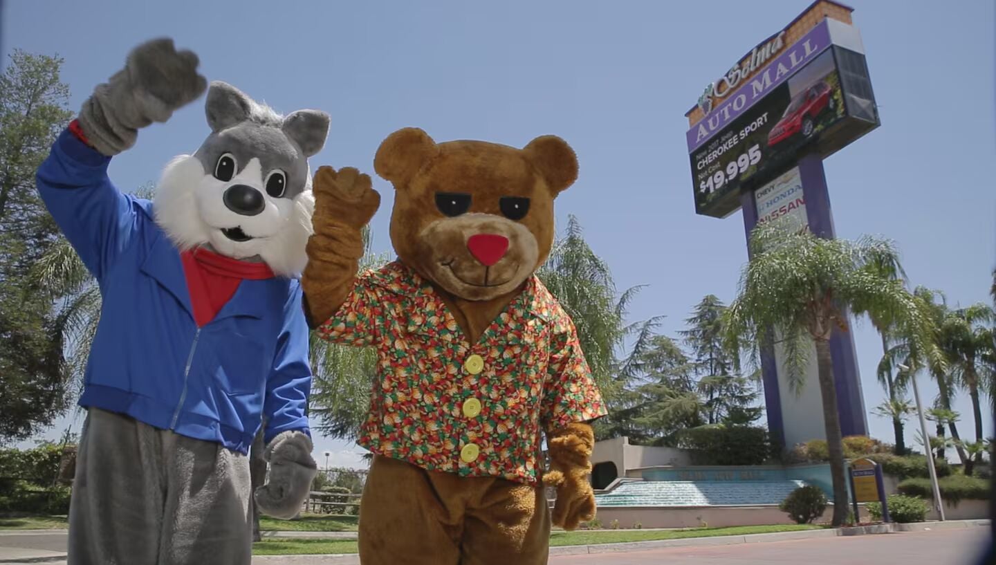 A person in a gray wolf mascot costume and a person in a brown bear mascot costume wave, with an auto mall sign and palm trees behind them.