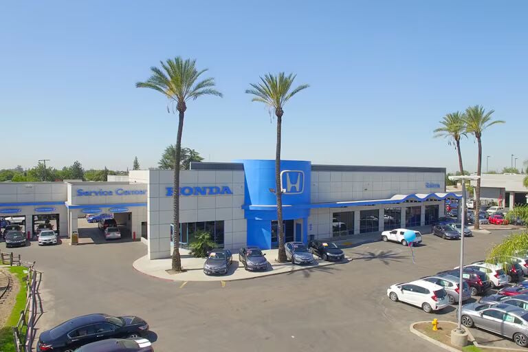 Exterior view of a Honda car dealership with several cars parked outside, palm trees in front, and a clear blue sky overhead.